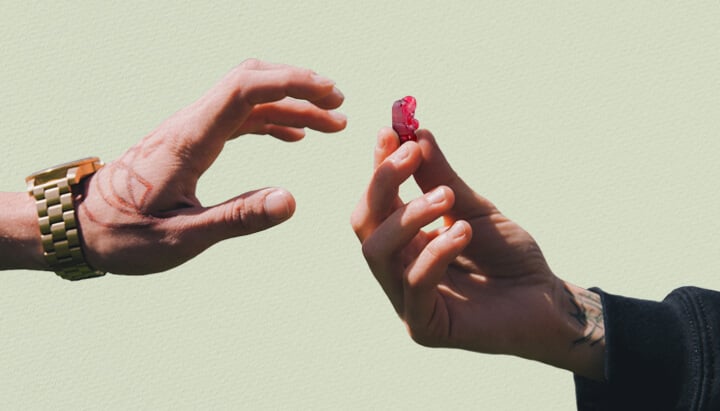 Zwei Hände greifen nacheinander, eine davon tätowiert, und halten ein rotes THC-Gummibärchen.